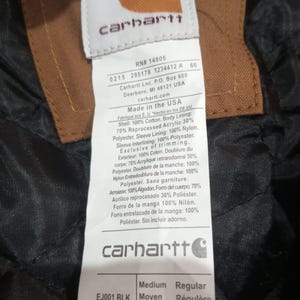 K&ouml;nnte beinhalten: Nahaufnahme eines Carhartt-Etiketts mit dem Markenlogo und Produktinformationen. Das Etikett ist wei&szlig; mit schwarzem Text, der Hintergrund ist ein dunkler, strukturierter Stoff. Das Etikett enth&auml;lt den Text "Made in the USA".