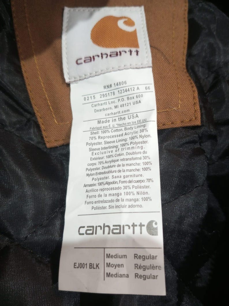 K&ouml;nnte beinhalten: Nahaufnahme eines Carhartt-Etiketts mit dem Markenlogo und Produktinformationen. Das Etikett ist wei&szlig; mit schwarzem Text, der Hintergrund ist ein dunkler, strukturierter Stoff. Das Etikett enth&auml;lt den Text "Made in the USA".