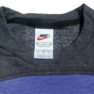 Op de afbeelding: Een close-up van een Nike sweatshirt. Het bovenste gedeelte is donkergrijs, met een zwarte kraag. Het onderste gedeelte is blauw. Een wit label met het Nike-logo en maat informatie is zichtbaar.