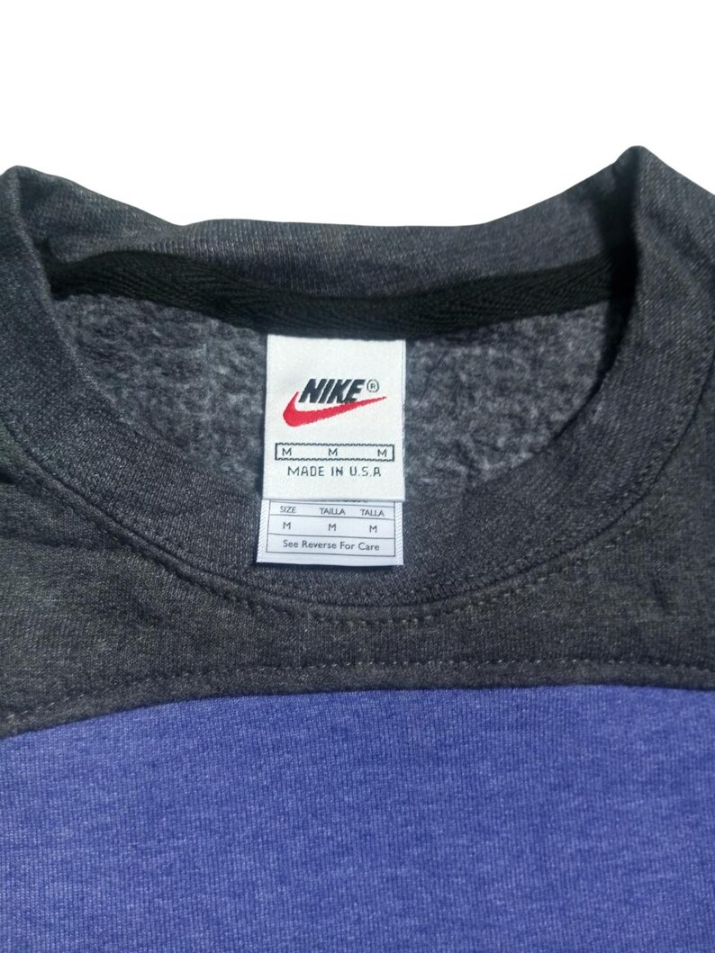 Op de afbeelding: Een close-up van een Nike sweatshirt. Het bovenste gedeelte is donkergrijs, met een zwarte kraag. Het onderste gedeelte is blauw. Een wit label met het Nike-logo en maat informatie is zichtbaar.