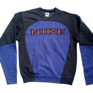 Op de afbeelding: Een grijs, blauw en paars Nike sweatshirt. Het sweatshirt heeft een grote paarse ovaal op de voorkant met het woord "NIKE" in bruine letters. De mouwen zijn blauw en de body is grijs.