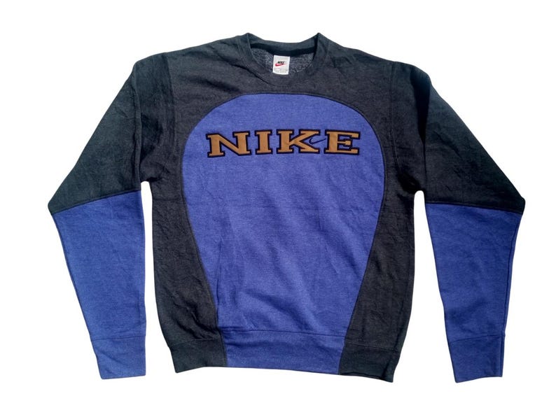 Op de afbeelding: Een grijs, blauw en paars Nike sweatshirt. Het sweatshirt heeft een grote paarse ovaal op de voorkant met het woord "NIKE" in bruine letters. De mouwen zijn blauw en de body is grijs.