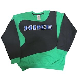 May include: Green and black Nike sweatshirt. The Nike logo in blue with a white outline is displayed prominently. The sleeves and upper section are black, with the lower section in green.