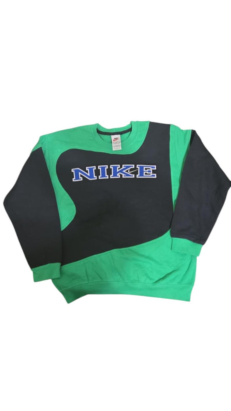 May include: Green and black Nike sweatshirt. The Nike logo in blue with a white outline is displayed prominently. The sleeves and upper section are black, with the lower section in green.