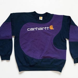 Puede incluir: Sudadera Carhartt azul marino y morada. La sudadera presenta un panel central morado con el logo de Carhartt en blanco y un peque&ntilde;o gr&aacute;fico naranja. Las mangas y la parte superior del cuerpo son azul marino con detalles morados.