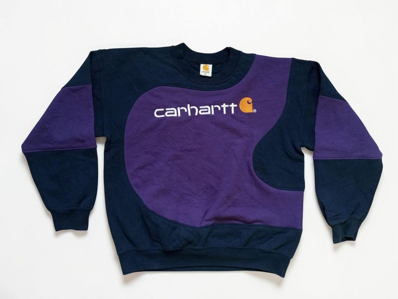 Puede incluir: Sudadera Carhartt azul marino y morada. La sudadera presenta un panel central morado con el logo de Carhartt en blanco y un peque&ntilde;o gr&aacute;fico naranja. Las mangas y la parte superior del cuerpo son azul marino con detalles morados.