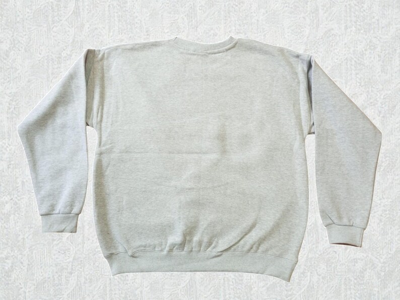 May include: A light gray sweatshirt with a crew neck and long sleeves. The sweatshirt is made of a soft material and has ribbed cuffs and hem. The garment is laid flat on a textured surface.