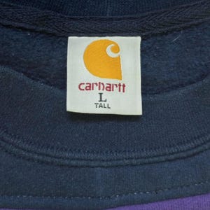 Puede incluir: Primer plano de una etiqueta de sudadera Carhartt. La etiqueta es blanca con el logotipo de Carhartt en naranja y el nombre de la marca en rojo. La etiqueta tambi&eacute;n indica la talla Large y Tall. La sudadera es azul marino y morada.