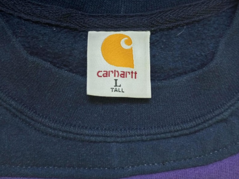 Puede incluir: Primer plano de una etiqueta de sudadera Carhartt. La etiqueta es blanca con el logotipo de Carhartt en naranja y el nombre de la marca en rojo. La etiqueta tambi&eacute;n indica la talla Large y Tall. La sudadera es azul marino y morada.