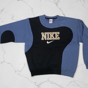 May include: A blue, black, and tan Nike sweatshirt. The sweatshirt has a black center panel with the Nike logo in tan block letters and a small white swoosh. The sleeves and upper portion are blue.