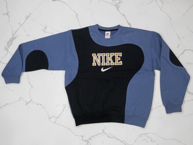 May include: A blue, black, and tan Nike sweatshirt. The sweatshirt has a black center panel with the Nike logo in tan block letters and a small white swoosh. The sleeves and upper portion are blue.