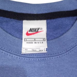 May include: Close-up of a blue crew neck sweatshirt with a black collar. A white tag with the Nike logo and "MADE IN U.S.A." is visible. The tag also includes size information.