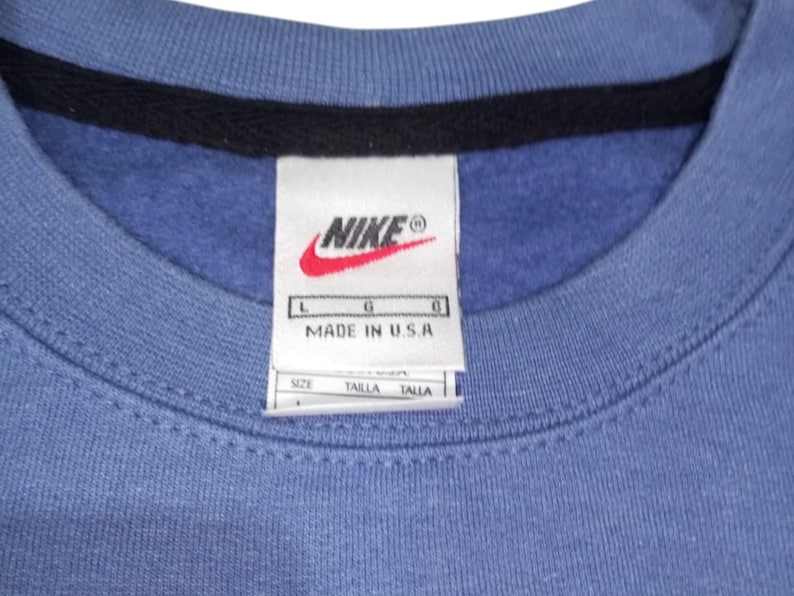 May include: Close-up of a blue crew neck sweatshirt with a black collar. A white tag with the Nike logo and "MADE IN U.S.A." is visible. The tag also includes size information.