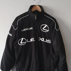 May include: A black zip-up jacket featuring the white Lexus logo and text embroidered on the chest and sleeves. The jacket has a high collar, long sleeves with white piping details, and a full-length front zip closure with a silver metal pull.