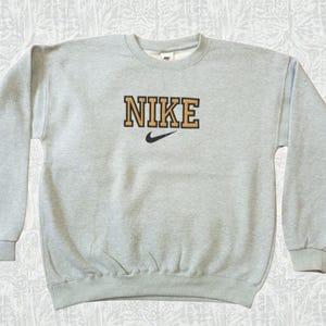May include: A light gray crew neck sweatshirt with the word "NIKE" in a gold and black block letter design across the chest. A black Nike swoosh logo is below the word. The sweatshirt has long sleeves and ribbed cuffs and hem.