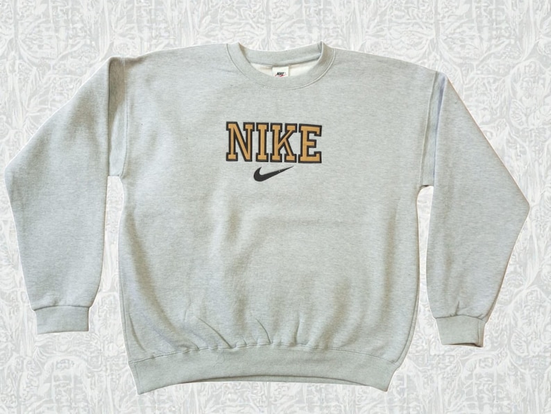 May include: A light gray crew neck sweatshirt with the word "NIKE" in a gold and black block letter design across the chest. A black Nike swoosh logo is below the word. The sweatshirt has long sleeves and ribbed cuffs and hem.