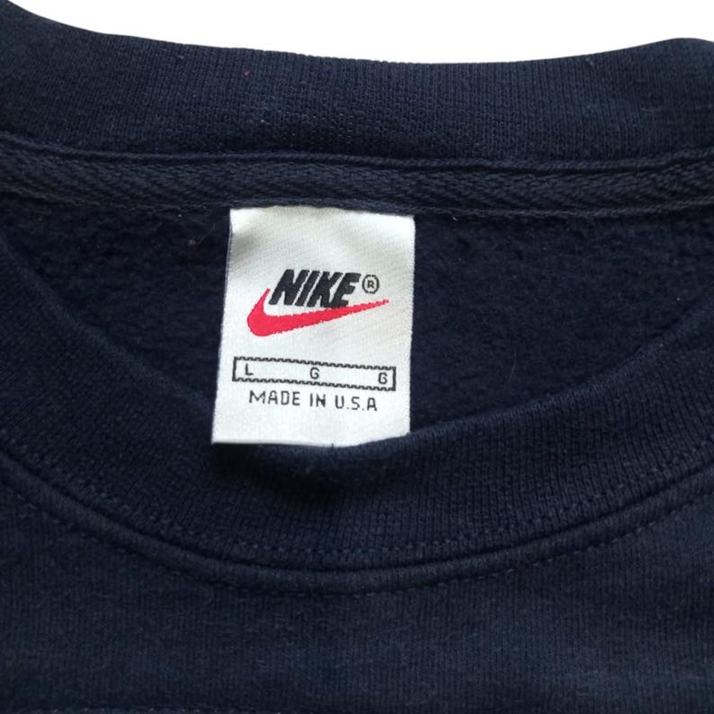 K&ouml;nnte beinhalten: Nahaufnahme eines marineblauen Sweatshirts mit einem wei&szlig;en Etikett. Das Etikett zeigt das Nike-Logo in Rot und Wei&szlig;, mit dem Markennamen und dem Text "MADE IN U.S.A." Das Sweatshirt hat einen Rundhalsausschnitt.