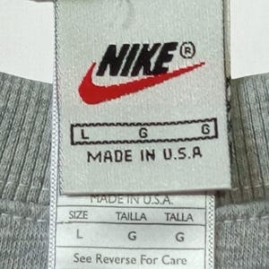 May include: Close-up of a gray crew neck shirt tag. The tag features the Nike logo in black and red, with the text "MADE IN U.S.A." and size information. The shirt is a size L.