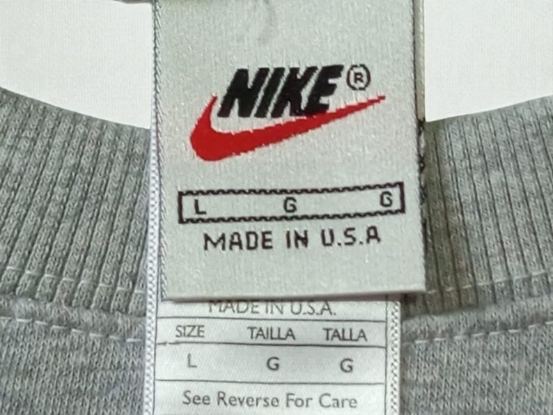 May include: Close-up of a gray crew neck shirt tag. The tag features the Nike logo in black and red, with the text "MADE IN U.S.A." and size information. The shirt is a size L.