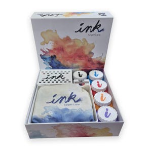 Könnte beinhalten: Eine weiße Box mit der Aufschrift "ink" und farbenfrohen Aquarell-Illustrationen. Im Inneren befinden sich Stempelkissen, Stempel und ein Stoffbeutel, alle mit "ink" und "Kasper Lapp" versehen. Das Set enthält Tinte in verschiedenen Farben.