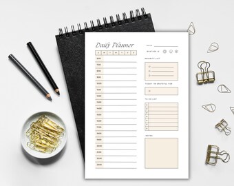 May include: A white daily planner with a black spiral binding, featuring a schedule from 6:00 to 23:00. Includes sections for the date, weather, priority list, gratitude, to-do list, and notes. Gold paper clips and pencils are also visible.