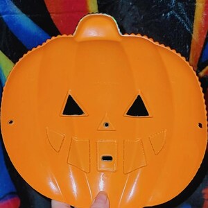 May include: An orange plastic jack-o'-lantern face with triangular eyes, a small nose, and a rectangular mouth. The pumpkin has a scalloped edge and a handle at the top. A vintage Halloween decoration.
