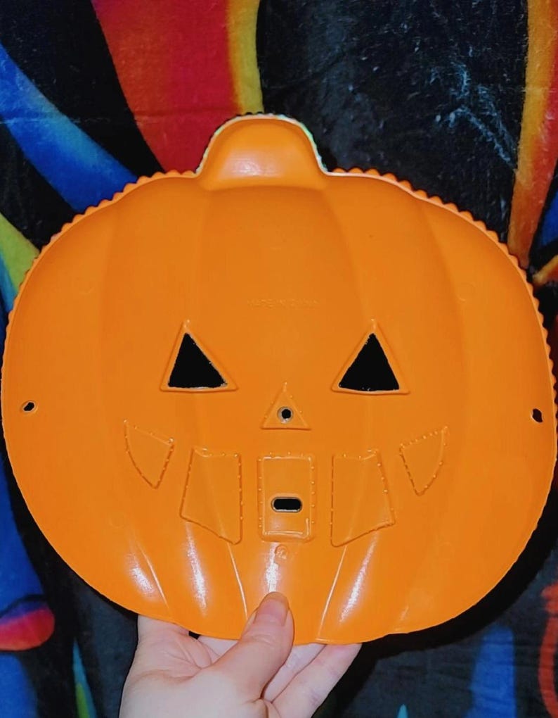 May include: An orange plastic jack-o'-lantern face with triangular eyes, a small nose, and a rectangular mouth. The pumpkin has a scalloped edge and a handle at the top. A vintage Halloween decoration.