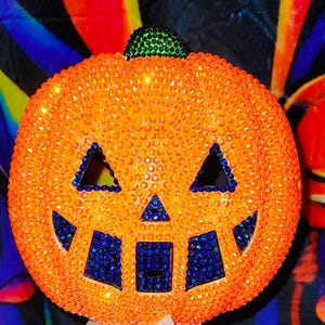 May include: An orange pumpkin decoration covered in small gems, with a green stem and blue accents. The pumpkin has triangular eyes and a square mouth, all outlined with blue gems. A festive Halloween decoration.