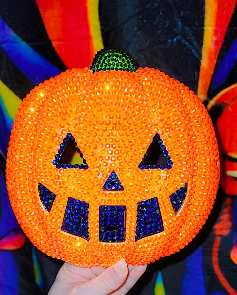 May include: An orange pumpkin decoration covered in small gems, with a green stem and blue accents. The pumpkin has triangular eyes and a square mouth, all outlined with blue gems. A festive Halloween decoration.