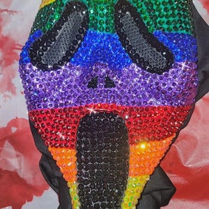 May include: A ghost-style mask covered in vibrant, multi-colored rhinestones arranged in horizontal stripes of orange, yellow, green, blue, purple, and red. The eye sockets and the elongated, open mouth are filled with solid black rhinestones.