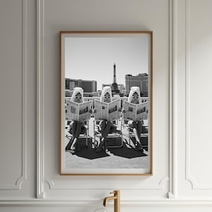 May include: Black and white framed print featuring three figures in sunglasses and headscarves reading newspapers. The Eiffel Tower and city buildings are visible in the background. The print is displayed above a white bathtub with a gold faucet.
