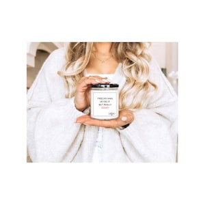 May include: A white jar candle with a black lid. The label reads "FREE MY MAN HE DID IT BUT REALLY IDGAF". The candle is held in a person's hand. The person is wearing a white sweater.