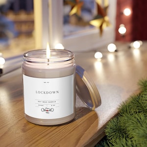 May include: A lit soy wax candle in a clear glass jar with a metal lid. The label reads "LOCKDOWN" and "SOY WAX CANDLE". The candle is 3 inches tall and contains 9 oz of wax. The jar sits on a wooden surface.