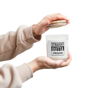 May include: A white candle in a frosted glass jar with a wooden lid. The label on the jar reads "STRAIGHT OUTTA PRISON" in bold, black capital letters. The candle is held in front of a white background.