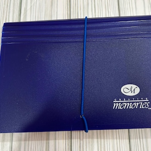 May include: A dark blue photo album with a blue elastic band closure. The album has a textured surface and the words "CREATIVE memories." in white. The album is lying on a light wood surface.
