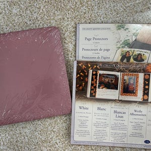 Puede incluir: Un álbum de fotos rosa polvoriento envuelto en plástico, junto a un paquete de protectores de páginas. Los protectores de páginas están etiquetados como "Page Protectors" y "The Creative Memories Collection". El paquete también presenta calabazas.