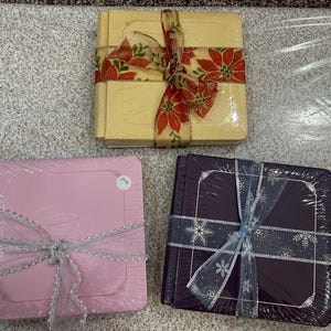 May include: Three wrapped square gift sets. One is yellow with a floral ribbon, one is pink with a silver ribbon, and one is dark purple with a snowflake ribbon. All are wrapped in clear plastic.
