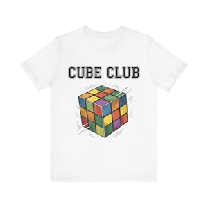 May include: A white short-sleeve t-shirt featuring the text CUBE CLUB in a dark grey collegiate font above a graphic of a multi-colored puzzle cube. The cube design includes motion lines to suggest rotation and features red, blue, yellow, and green.