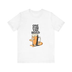 May include: A white short-sleeve t-shirt featuring a graphic of a beer mug filled with golden liquid and white foam. Below the mug is an orange road illustration. The text "ONE FOR THE ROAD" is printed in black above the beer mug graphic.