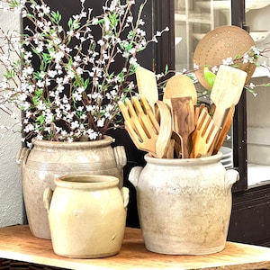 May include: A kitchen display with three ceramic pots and wooden utensils. The largest pot contains wooden spoons and spatulas. Two other pots hold white flowering branches. The beige pots sit on a wooden shelf. The scene has a rustic feel.