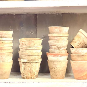 May include: A collection of terracotta flower pots stacked on a shelf. The pots vary in size and show signs of weathering, with some displaying a reddish-brown hue and others a lighter, aged appearance. The shelf is painted white.