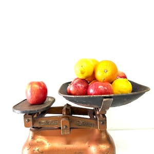May include: A vintage copper-colored metal balance scale with a dark iron tray and a small platform. The scale features the embossed text W & T AVERY LTD. A single red apple sits on the small platform, while a collection of red apples, yellow lemons, and an orange rests in the large iron tray.