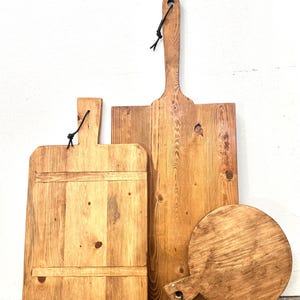 May include: A collection of wooden cutting boards in various shapes and sizes. The boards are made of light-colored wood with visible grain and knots. Each board has a handle and a black leather loop for hanging. The boards range in size from a small circle to a large rectangle.