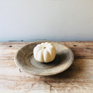 May include: A small, white pumpkin with a golden stem sits in a rustic, weathered wooden bowl. The bowl is round and has a wide rim. The pumpkin is the focal point, with the bowl and wooden surface providing a neutral backdrop.