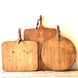 Puede incluir: Tres tablas de cortar de madera de diferentes formas. Las tablas son rectangulares, cuadradas y redondas, con un acabado natural de vetas de madera. Cada tabla tiene un asa con un lazo de cuero negro para colgar. Se exhiben sobre una superficie de madera.