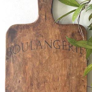 May include: A rectangular wooden bread board with a handle at the top, featuring a dark brown, weathered grain. The text BOULANGERIE is printed in black near the top, with AU BON PAIN and N 12 printed in black toward the bottom of the board.