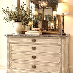 Può includere: Una cassettiera in legno grigio chiaro con quattro cassetti, ognuno dotato di una maniglia in metallo scuro. La cassettiera ha un piano in marmo grigio scuro. Sulla superficie poggiano un vaso in ceramica beige con rami, due libri e una lampada.
