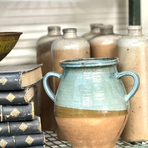 May include: A two-handled ceramic vase with a light blue upper section, a brown band, and a tan lower section. The vase is displayed on a metal shelf with other antique bottles and books.