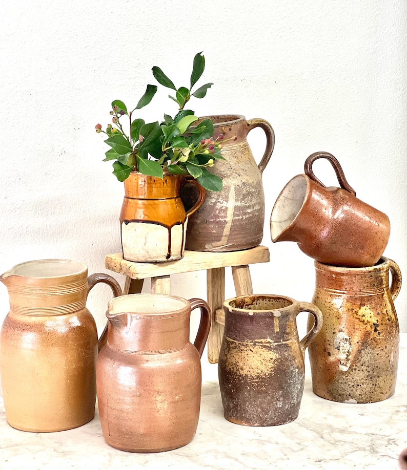 May include: A collection of six vintage brown pottery jugs and pitchers, some with a white glaze, on a wooden stool. The jugs are various sizes and shapes, with some having handles.