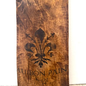 Puede incluir: Una tabla de madera rectangular con un acabado teñido en marrón oscuro y veta de madera visible. Un símbolo de flor de lis negro está impreso en el centro. Debajo del símbolo, el texto dice AU BON PAIN y N° 12 en letras negras.